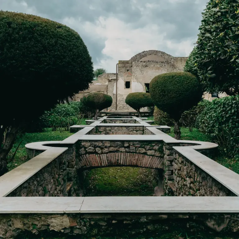 View of the Euripus water channel in the garden of the House of Julia Felix, Pompeii, framed by manicured trees and ancient stone walls under a cloudy sky.